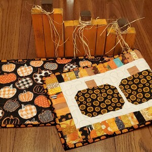 May include: Wooden orange pumpkin decorations with brown stems and raffia, alongside two quilted placemats. One placemat features a black background with orange and white pumpkins, while the other has a white background with sunflower-filled pumpkin shapes.
