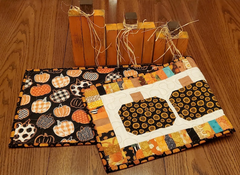 May include: Wooden orange pumpkin decorations with brown stems and raffia, alongside two quilted placemats. One placemat features a black background with orange and white pumpkins, while the other has a white background with sunflower-filled pumpkin shapes.