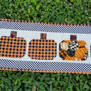 May include: A rectangular quilted table runner featuring a black and white checkered border and five pumpkin shapes appliqued in a row. Four pumpkins have a black and orange diamond pattern, while the fourth pumpkin features a multi-colored print.