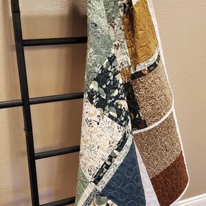 May include: A patchwork quilt featuring various fabric squares in shades of sage green, navy blue, mustard yellow, and brown with floral and botanical patterns. The quilt has white binding and is draped over a black wooden ladder next to a brass lantern.