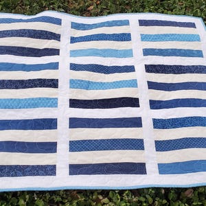 May include: A rectangular quilt with a white border and a light blue binding. The quilt features alternating horizontal stripes of various shades of blue and off-white. The quilt is approximately 50 inches wide and 60 inches long.