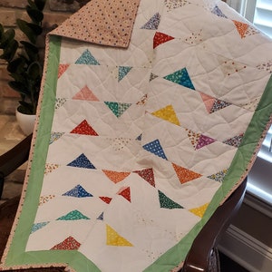 May include: A handmade quilt with a white background and colorful triangle patterns. The triangles are in various colors, including red, blue, yellow, and green. The quilt has a green border and a patterned backing.