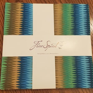 May include: A stack of fabric squares with a repeating geometric pattern in shades of blue, green, and gold. The fabric is wrapped with a white label featuring the text "Free Spirit" in a script font.