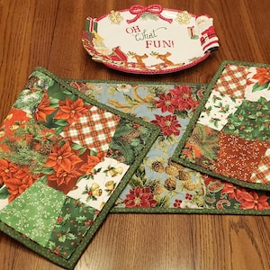 May include: A rectangular quilted table runner featuring a patchwork design of poinsettias, pine cones, and plaid patterns in red, green, and white, accompanied by a white ceramic serving dish with a red bow and the text OH what FUN!