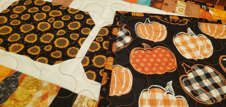 May include: Close-up of a quilt with various fabric patterns. One section features sunflowers on a black background. Another shows orange pumpkins with white outlines on a black background. The quilt has white and orange accents.