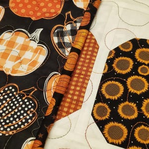 May include: A patchwork quilt with various autumn-themed patterns. The quilt features pumpkins in orange, black, and white, with patterns like polka dots, stripes, and gingham. Another section has sunflowers on a black background.