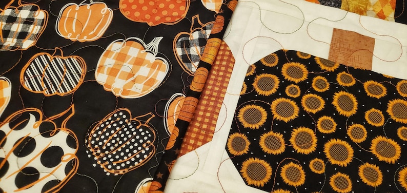 May include: A patchwork quilt with various autumn-themed patterns. The quilt features pumpkins in orange, black, and white, with patterns like polka dots, stripes, and gingham. Another section has sunflowers on a black background.