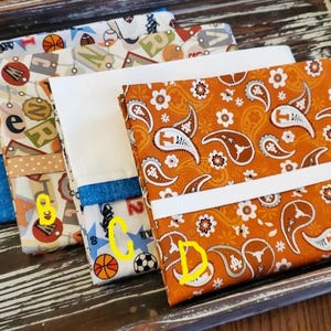 May include: Four fabric items with different patterns and colors are displayed on a wooden tray. The items include sports-themed prints, a white fabric with a blue border, and an orange paisley design. The tray has black handles.
