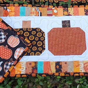 May include: A handmade, quilted table runner and matching placemat featuring a fall harvest theme. The runner has three pumpkin shapes in the center, with a white background and a border of orange, brown, and black fabrics. The placemat has a black background with various pumpkin designs.