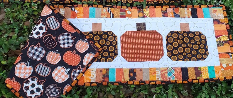 May include: A handmade, quilted table runner and matching placemat featuring a fall harvest theme. The runner has three pumpkin shapes in the center, with a white background and a border of orange, brown, and black fabrics. The placemat has a black background with various pumpkin designs.