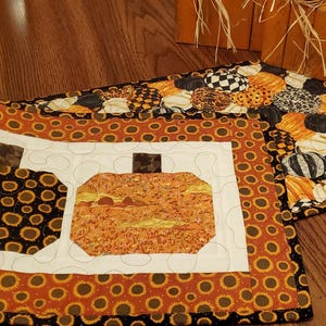 May include: A quilted table runner with a fall theme. The runner features two pumpkin shapes on a white background, surrounded by a border of orange sunflowers. The runner is accented with a black border. A second runner with a pumpkin pattern is also visible.