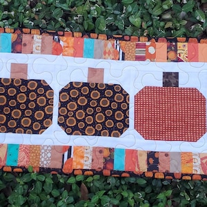 May include: A rectangular quilted table runner featuring five pumpkin shapes. Four pumpkins have a black and gold circular pattern, while the center pumpkin is solid orange. The runner has a white background with a colorful patchwork border.