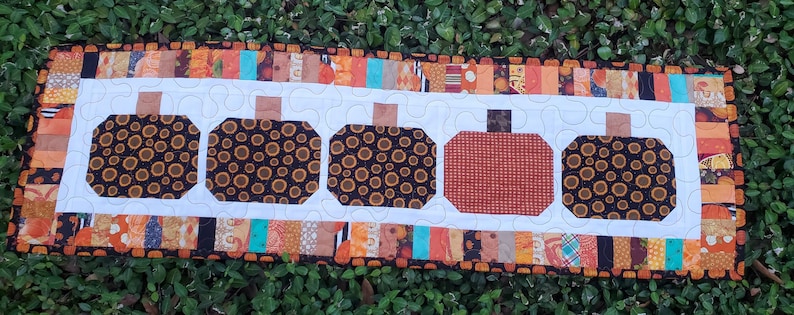 May include: A rectangular quilted table runner featuring five pumpkin shapes. Four pumpkins have a black and gold circular pattern, while the center pumpkin is solid orange. The runner has a white background with a colorful patchwork border.