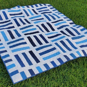 May include: A blue and white quilt with a geometric pattern of horizontal and vertical stripes. The quilt features various shades of blue, including navy, light blue, and sky blue, set against a white background. The quilt is displayed on a bed of green grass.