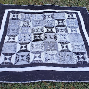 May include: A handmade quilt with a black and white color scheme. The quilt features a central pattern of squares and geometric shapes in various shades of gray, black, and white. A white border frames the central design, and a black border with a decorative stitch surrounds the quilt.