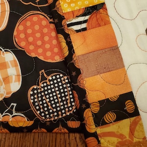 May include: A patchwork quilt featuring autumn-themed patterns. The quilt showcases pumpkins in various designs, including polka dots and gingham, set against a black background. Adjacent to the pumpkin pattern is a section with sunflowers.