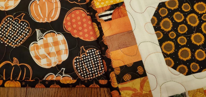 May include: A patchwork quilt featuring autumn-themed patterns. The quilt showcases pumpkins in various designs, including polka dots and gingham, set against a black background. Adjacent to the pumpkin pattern is a section with sunflowers.