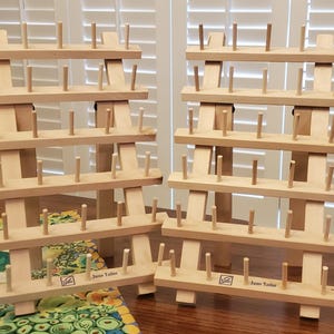 May include: Two wooden thread spool holders with multiple rows of pegs. Each holder has a rectangular base and is labeled "Jane Taylor". The holders are made of light-colored wood and are designed to organize spools of thread.