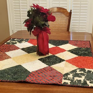 May include: A wooden dining table is set with a decorative quilted table runner. The runner features a diamond pattern in red, green, cream, and black, with a deer silhouette design. A vase of red flowers sits in the center.