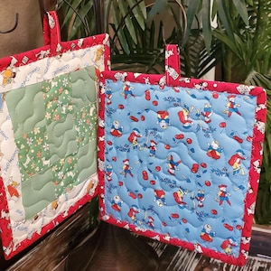 May include: Two quilted pot holders with red trim. One is green with a floral pattern and the text "Sweet Dreams" and "My Moods". The other is blue with a pattern of children in red and white outfits and red cherries.