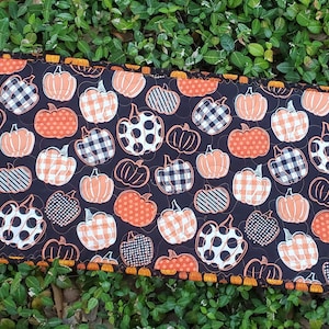 May include: A rectangular, quilted table runner featuring a repeating pattern of orange, white, and black pumpkins on a dark background. The pumpkins have various patterns, including polka dots and plaid. The runner is trimmed with an orange and brown zigzag edge.