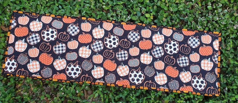 May include: A rectangular, quilted table runner featuring a repeating pattern of orange, white, and black pumpkins on a dark background. The pumpkins have various patterns, including polka dots and plaid. The runner is trimmed with an orange and brown zigzag edge.