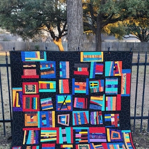May include: A colorful quilt with a black background and a patchwork design. The quilt features rectangular blocks in various colors, including red, blue, yellow, and green. The blocks are arranged in a grid pattern and are outlined with black stitching. The quilt is displayed outdoors.