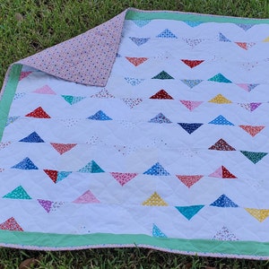 May include: A handmade quilt with a white background and rows of colorful triangle patterns. The triangles are in various colors and patterns, including red, blue, green, and yellow. The quilt has a green border and a floral patterned backing.