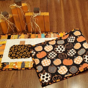 May include: A fall-themed display featuring three wooden pumpkin decorations and two quilted placemats. One placemat has a black background with orange and white pumpkins, while the other has a white center with a sunflower design.