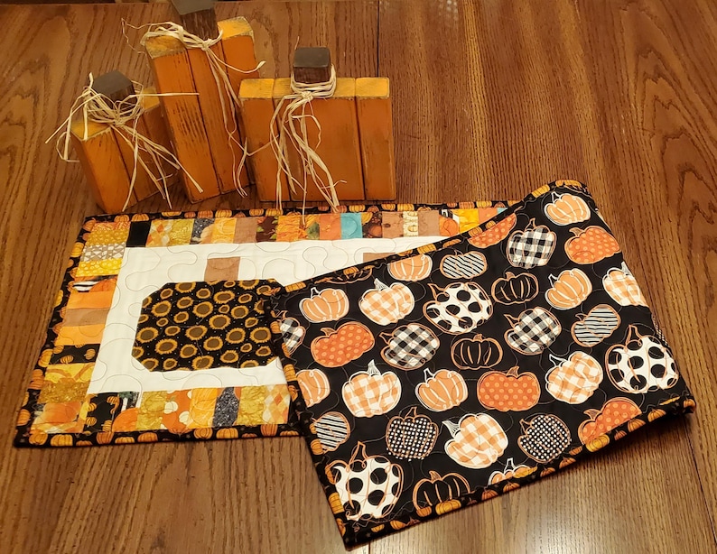May include: A fall-themed display featuring three wooden pumpkin decorations and two quilted placemats. One placemat has a black background with orange and white pumpkins, while the other has a white center with a sunflower design.