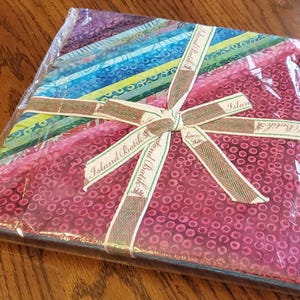 May include: A fabric bundle wrapped in clear plastic, tied with a ribbon. The fabric features a variety of colorful patterns, including stripes and circular designs. The ribbon is beige with the words "Island Batik" printed on it.