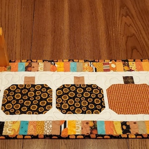May include: A fall-themed table runner with five pumpkin designs. The pumpkins are on a white background, with a colorful patchwork border. Wooden pumpkin decorations with raffia are on the left.