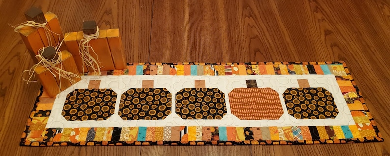 May include: A fall-themed table runner with five pumpkin designs. The pumpkins are on a white background, with a colorful patchwork border. Wooden pumpkin decorations with raffia are on the left.