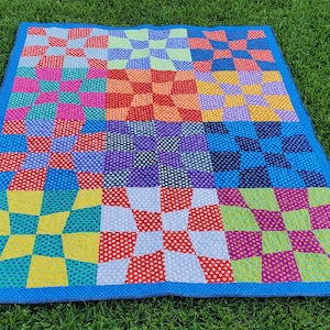 May include: A colorful patchwork quilt with a blue border rests on green grass. The quilt features a checkerboard pattern in various colors, including red, yellow, blue, purple, and green, with polka dot and solid squares.