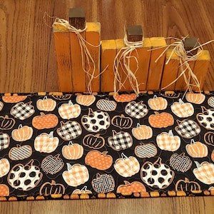 May include: A rectangular black table runner with a pumpkin pattern in orange, white, and black. Wooden pumpkin-shaped decorations with raffia accents sit on the wooden surface behind the runner. The runner is approximately 12 inches wide.