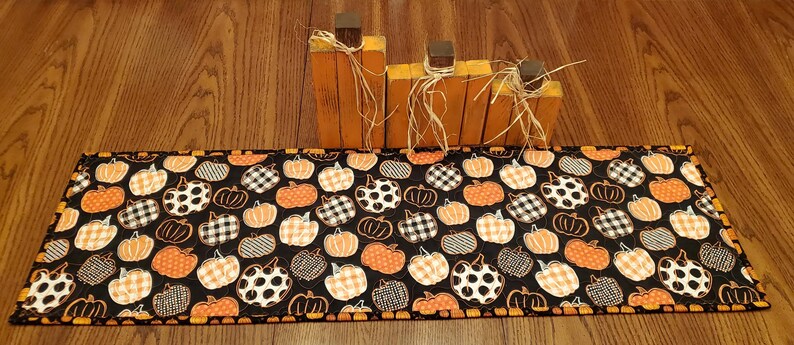 May include: A rectangular black table runner with a pumpkin pattern in orange, white, and black. Wooden pumpkin-shaped decorations with raffia accents sit on the wooden surface behind the runner. The runner is approximately 12 inches wide.
