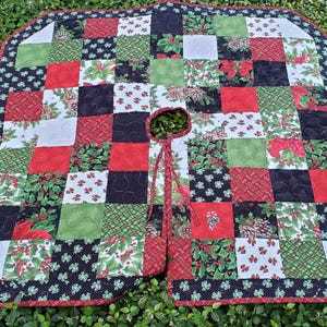 May include: A quilted Christmas tree skirt featuring a patchwork design of red, green, white, and black fabric squares. The pattern includes holly, poinsettia, and candy cane motifs with a central circular opening and two fabric ties.
