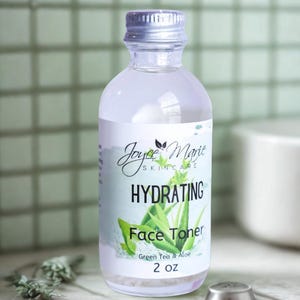 May include: A clear glass bottle with a silver lid containing a hydrating face toner. The label is white with black text that reads "Joyce Marie Skincare Hydrating Face Toner Green Tea & Aloe 2 oz".