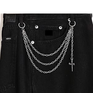 May include: Close-up of black denim jeans with a silver chain belt. The chain is composed of three strands and a small cross pendant. The jeans have a classic five-pocket design.