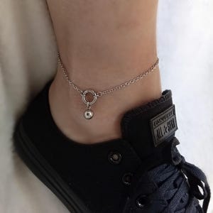 Op de afbeelding: Een zilveren enkelbandje met een kleine kogelhanger. De fijne ketting is bevestigd aan een cirkelvormige sluiting. Het enkelbandje wordt gedragen met een zwarte sneaker.