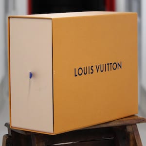May include: A rectangular Louis Vuitton gift box featuring a vibrant orange exterior with the brand name printed in dark blue lettering. The front panel is a light cream color and includes a small, thin blue ribbon pull tab for opening the box.