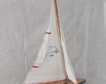 May include: A wooden sailboat model with a blue hull and a light brown deck. The white sail features a black dolphin illustration and the number 401. Red accents are on the sail. The model is resting on a stack of books.