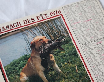 Op de afbeelding: Vintage kalender uit 1976 met een kleurenfoto van een gouden Labrador Retriever die een vogel in zijn bek heeft. De kalender heeft een rood-witte rand en de tekst "ALMANACH DES P.T.T. 1976". De kalenderpagina's zijn zichtbaar.