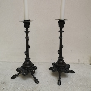 May include: Two black metal candle holders with ornate details and three legs. Each holder has a white candle on top.
