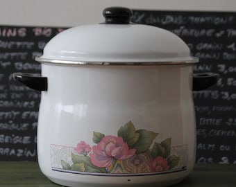 May include: White enamel cooking pot with a black lid and handles. The pot features a floral design with pink flowers and green leaves. A silver rim accents the top and bottom. The pot is sitting on a green surface.