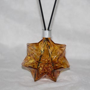 Pode incluir: Um frasco difusor de vidro âmbar em forma de estrela com um colar de metal prateado e duas varetas pretas finas que se estendem do topo. O vidro apresenta um design geométrico facetado que capta a luz, criando tons alaranjados e castanhos.