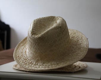 May include: A light-colored straw hat with a wide brim and a woven texture. The hat is resting on a white surface. The hat's crown is slightly dented, and the brim is curved.