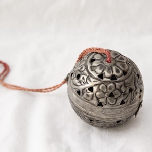 May include: A silver-coloured, spherical decorative object with floral cutouts and a braided cord. The object has a small red tassel and is likely used for decoration or as a hanging ornament. The background is a white fabric.