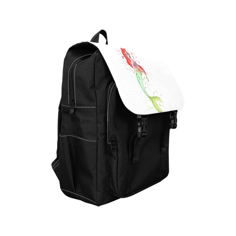 K&ouml;nnte beinhalten: Ein schwarzer Rucksack mit einer wei&szlig;en Klappe, die eine Aquarellillustration einer Meerjungfrau mit rotem Haar und gr&uuml;nem Schwanz zeigt. Der Rucksack verf&uuml;gt &uuml;ber schwarze Seitentaschen, schwarze Rei&szlig;verschl&uuml;sse und zwei schwarze Schnallen.