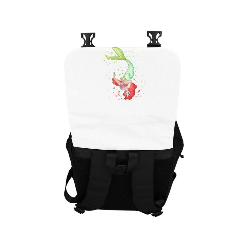 K&ouml;nnte beinhalten: Ein schwarzer Rucksack mit einer wei&szlig;en Klappe, die eine Illustration im Aquarellstil einer Meerjungfrau mit rotem Haar und gr&uuml;nem Schwanz zeigt. Der Rucksack verf&uuml;gt &uuml;ber verstellbare schwarze Schultergurte und zwei schwarze Kunststoffschnallen.